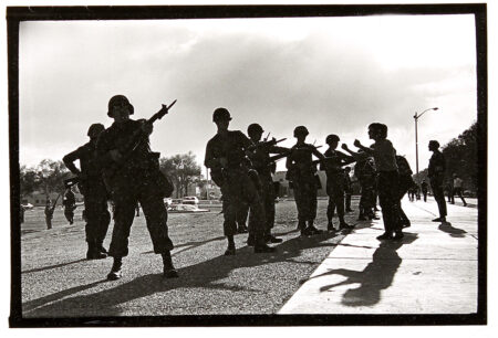 National Guardsmen stand in a group with bayonets out. A person stands in fornt of them with their arms extended..