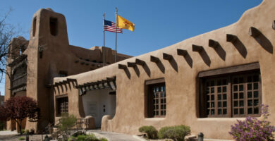 The entrance of the New Mexico Museum of Art's 1917 Plaza Building. A New Mexico State Flag and the US flag blow in the wind against a clear blue sky. The Pueblo Revival-style building is adobe with wooden vigas and window coverings.