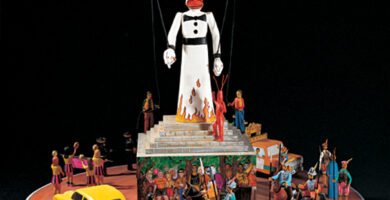 Colorful diorama of the puppet, Zozobra, and fiesta-goers surrounding him. The figurines are arranged on an orange circular base. A yellow car appears at the base of the Zozobra figure, along with people on horses.